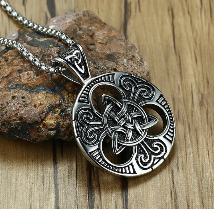 Lying against a rock is a round pendant that has a design in the middle of three interlocked triangles which then branch out over cut out pieces and then the outer circle. There are detailed patterns carved onto the whole thing and it is silver in colour.