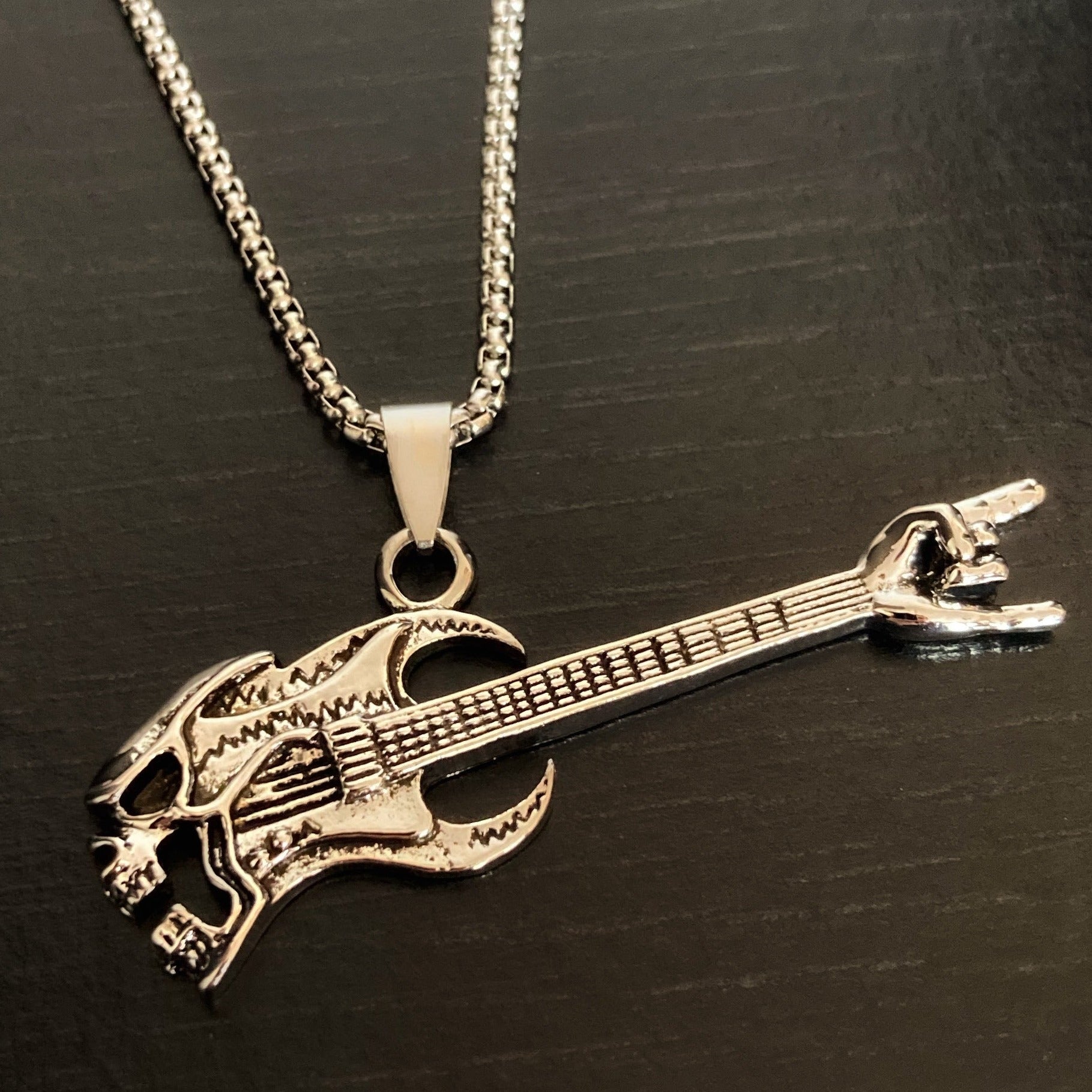 A necklace has a guitar shaped pendant hanging from it. These are both silver in colour with the guitar having detailed etchings on it of the strings etc and a snarling skulls face at one end.