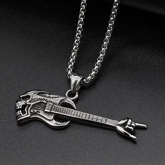 On a dark surface lies a pendant in the shape of a guitar. The main body of the guitar has a skull face and at the end of the strings there is a hand clenched with two fingers sticking out in a rocking out style.