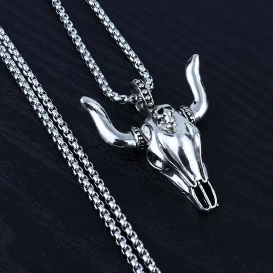 On a grainy black background sits a necklace that is silver in colour. The Pendant is in the shape of a bulls skull with horns and detailed markings. It is solid looking but light in weight to hold and the chain looks to be sturdy.