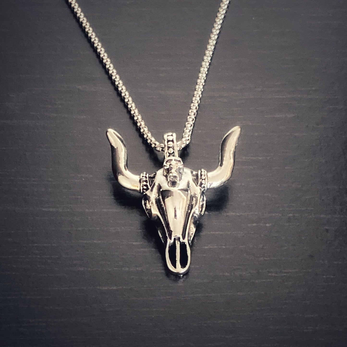 A close up view of a silver coloured bull skull shaped pendant. There are intricate markings on the skull and it is attached to a chain also the colour silver. 