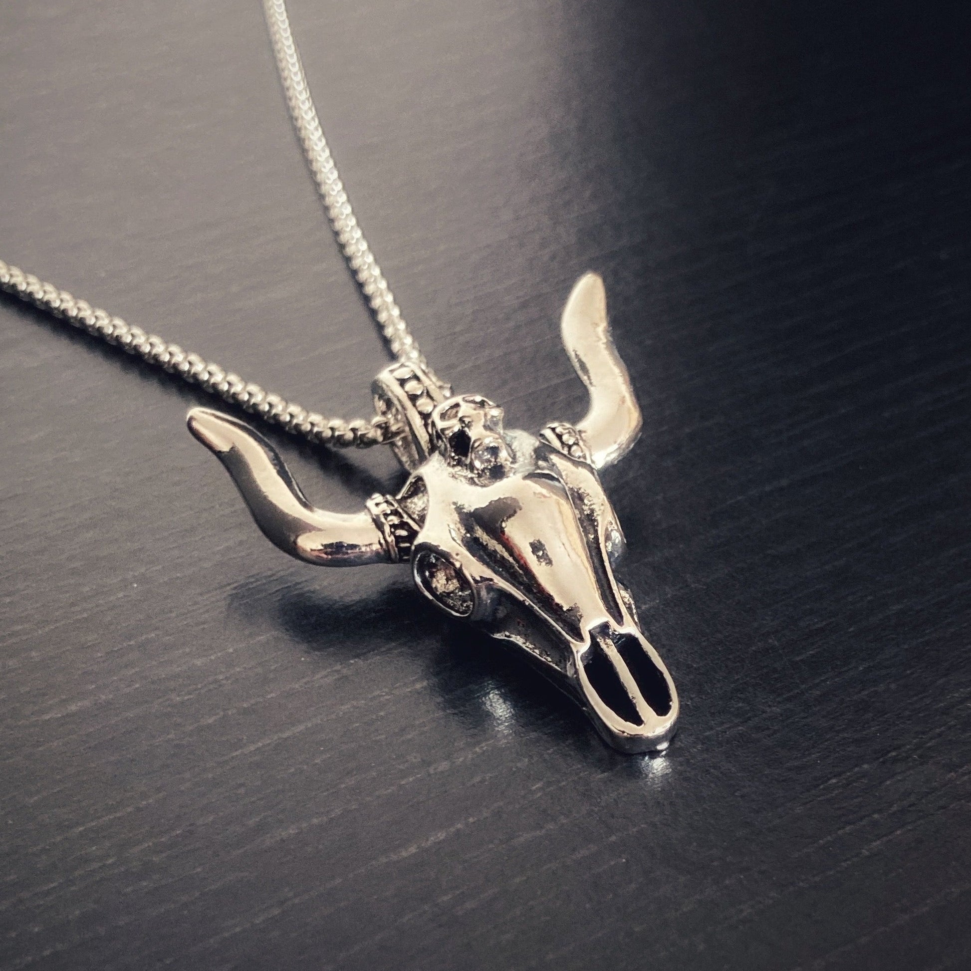 A side on view of a chain with a bulls skull shaped pendant attached to it by a round ring. Featuring eyes, nose and horns this item is silver in colour and made of stainless steel.