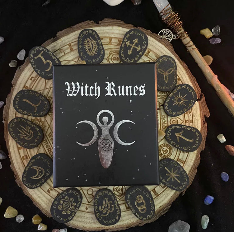 A round piece of wood holds thirteen black coloured wooden runes that are engraved in a gold cololur and are intricate in design. In the centre of the circle is a black box that says witch runes on it. 