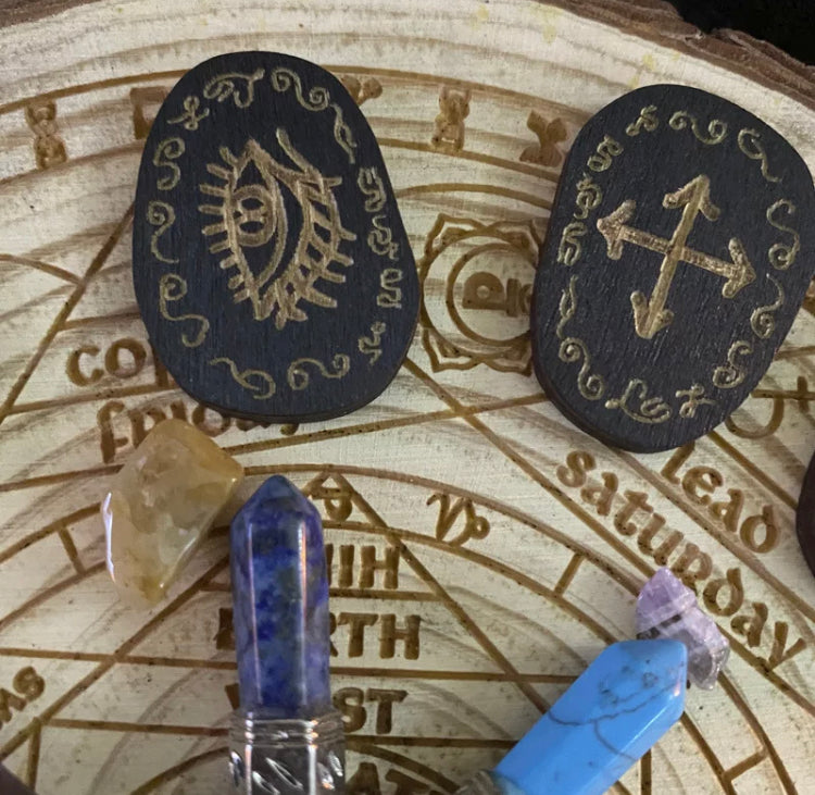 A close up view of two of the inscribed runes surrounded by crystals. You can see the beautiful detailed carvings on these items which are made of wood and are black in colour with the engravings looking a gold colour.