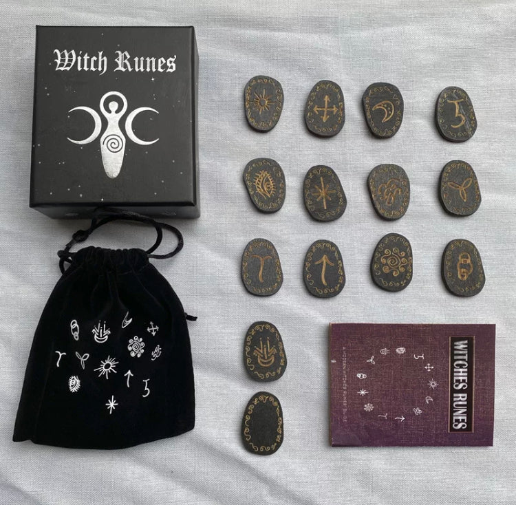 Layed out on a white cloth are all the components of a witches rune set. This includes the wooden runes themselves along with a black velvet bag, booklet and a box that it all comes in.