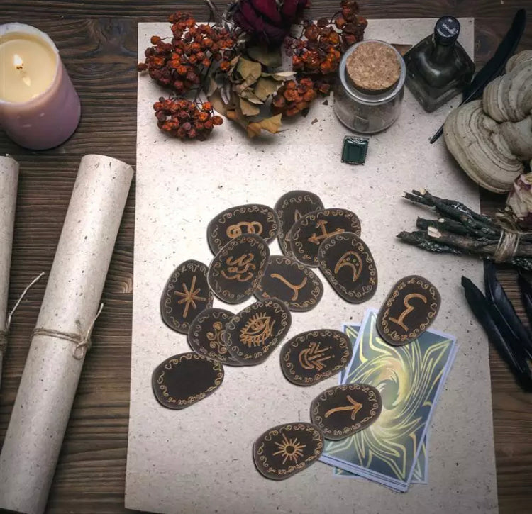 There are 14 wooden runes scattered onto a white background that also houses some dried plants and herbs and face down cards. 13 of these black wooden runes are inscribed with one being blank. They are oval in shape and very detailed.