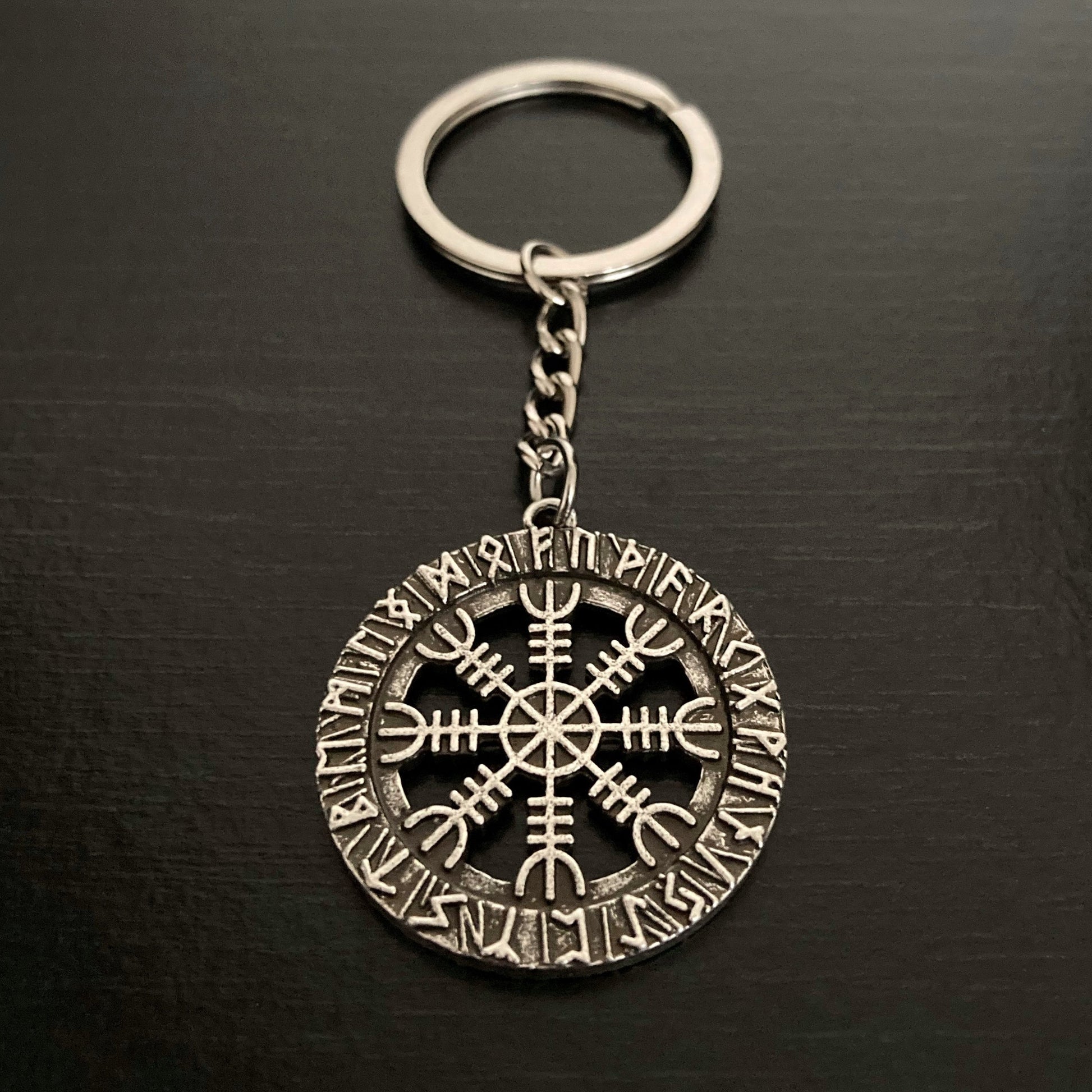 On a black background is a silver coloured keyring. Dangling from the ring by a small chain is a circular design featuring runic symbols round the outer part of the circle and a cut out helm of awe design in the centre.