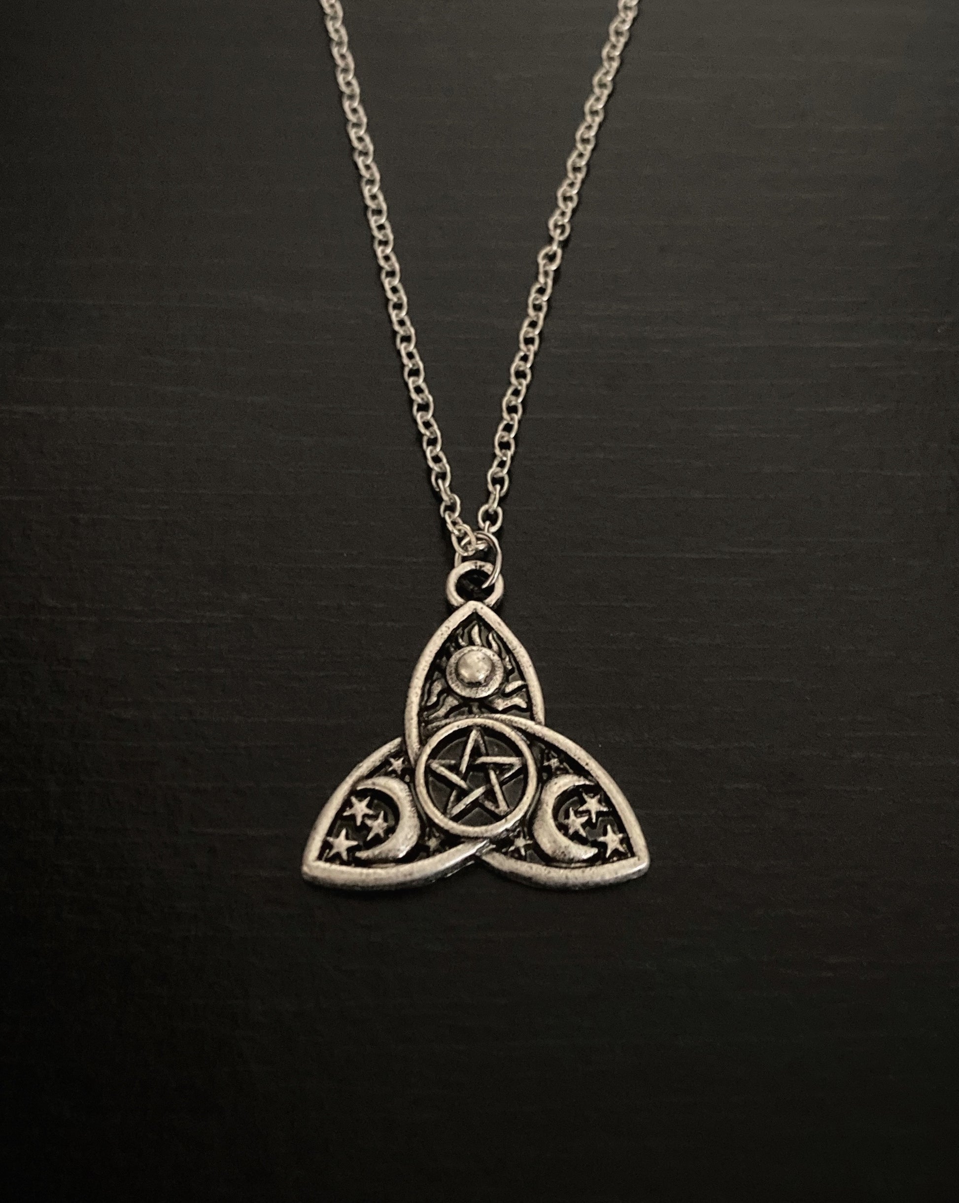 A silver coloured chain holds a pendant which has markings of a pentagram, moon, sun and stars covering the surface of a triple moon shape.