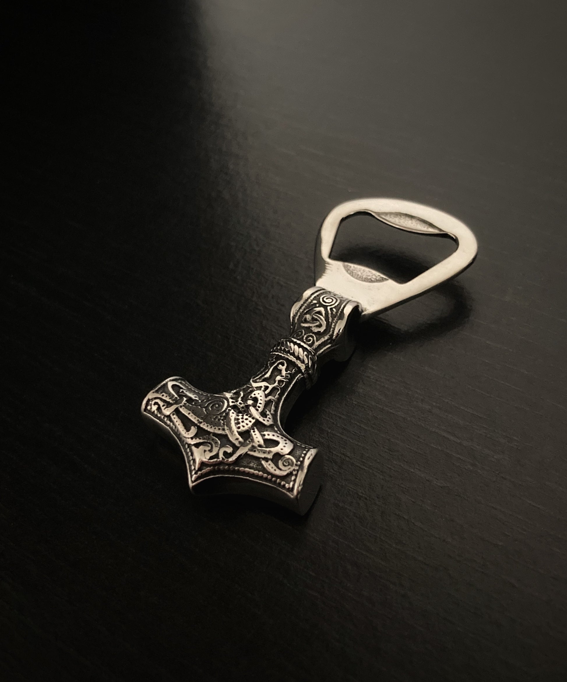 Lying on a black background is a bottle opener which is shaped like the hammer that Thor wielded. One end has a solid ring round it with a hole in the middle to place over a bottle cap for removal. The other end is solid with a design engraved on it.