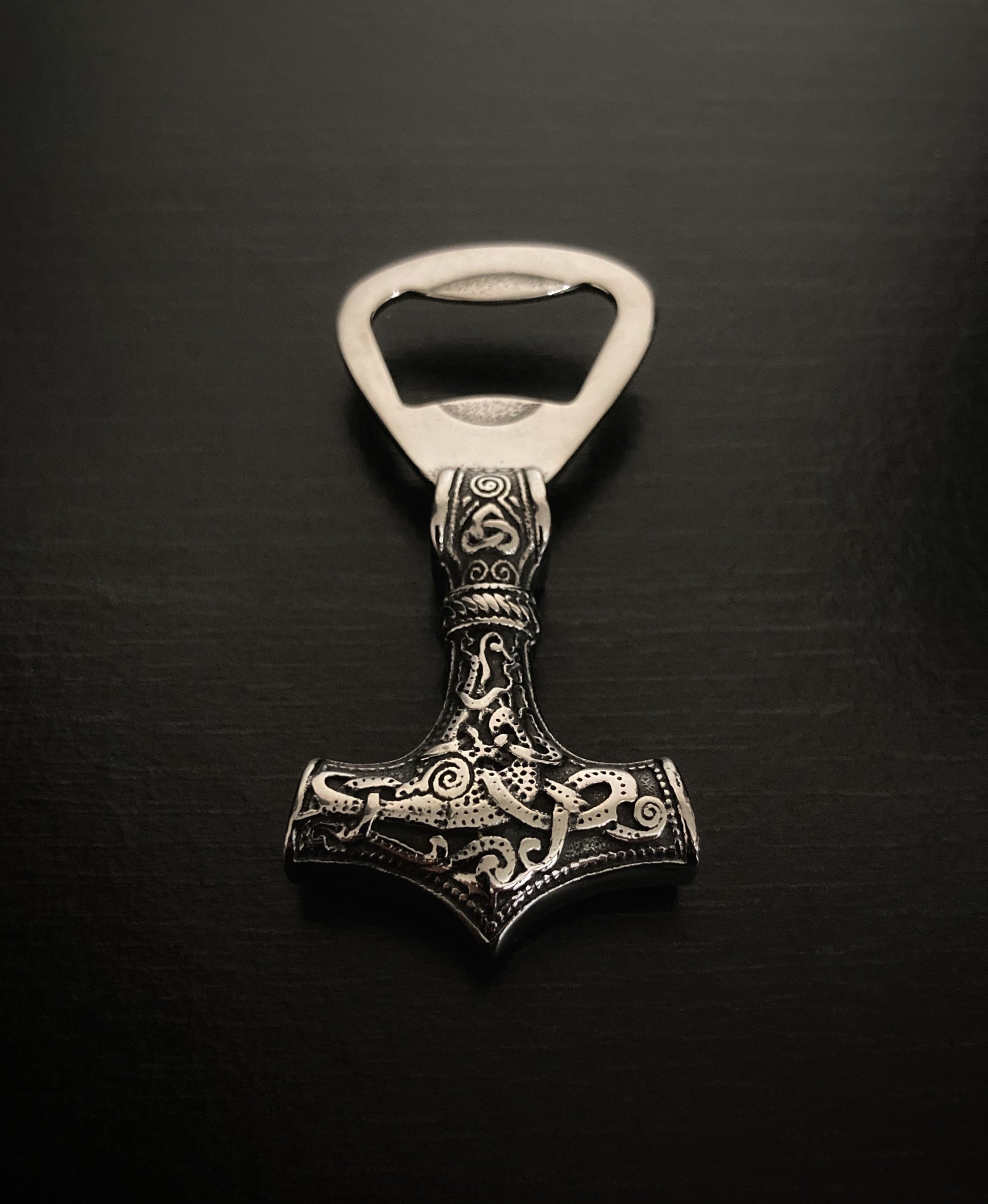 Sitting on a black plain surface is a solid weighty viking hammer bottle opener. It is heavy to hold in your hand and is made from stainless steel. 7cm in length this features a silver colour design running along one end and is double sided.