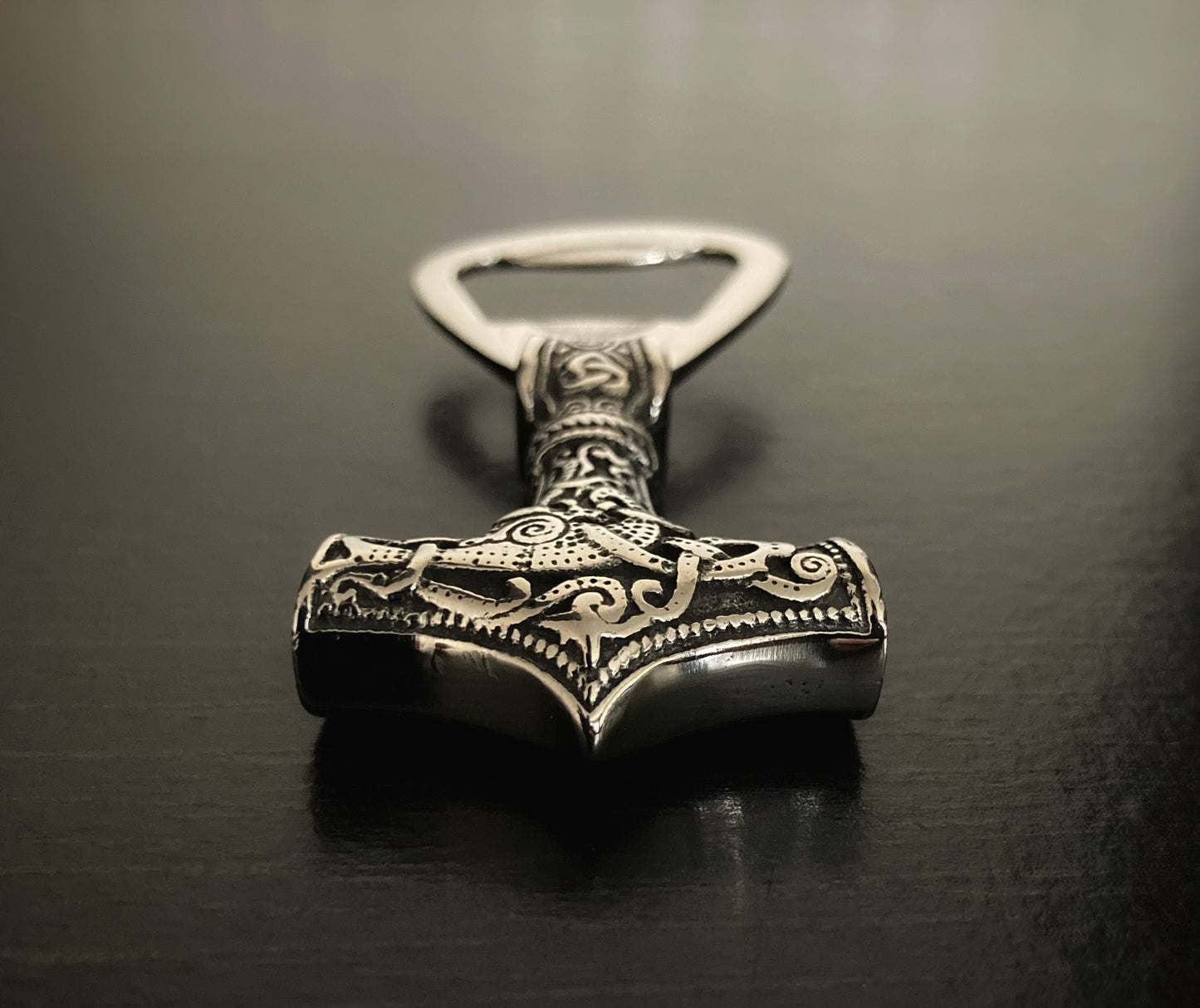 A close up of a stainless steel bottle opener in the shape of a viking hammer. You can see the design in detail that is engraved onto the item and if you touch it you can feel the pattern on your fingertips. 