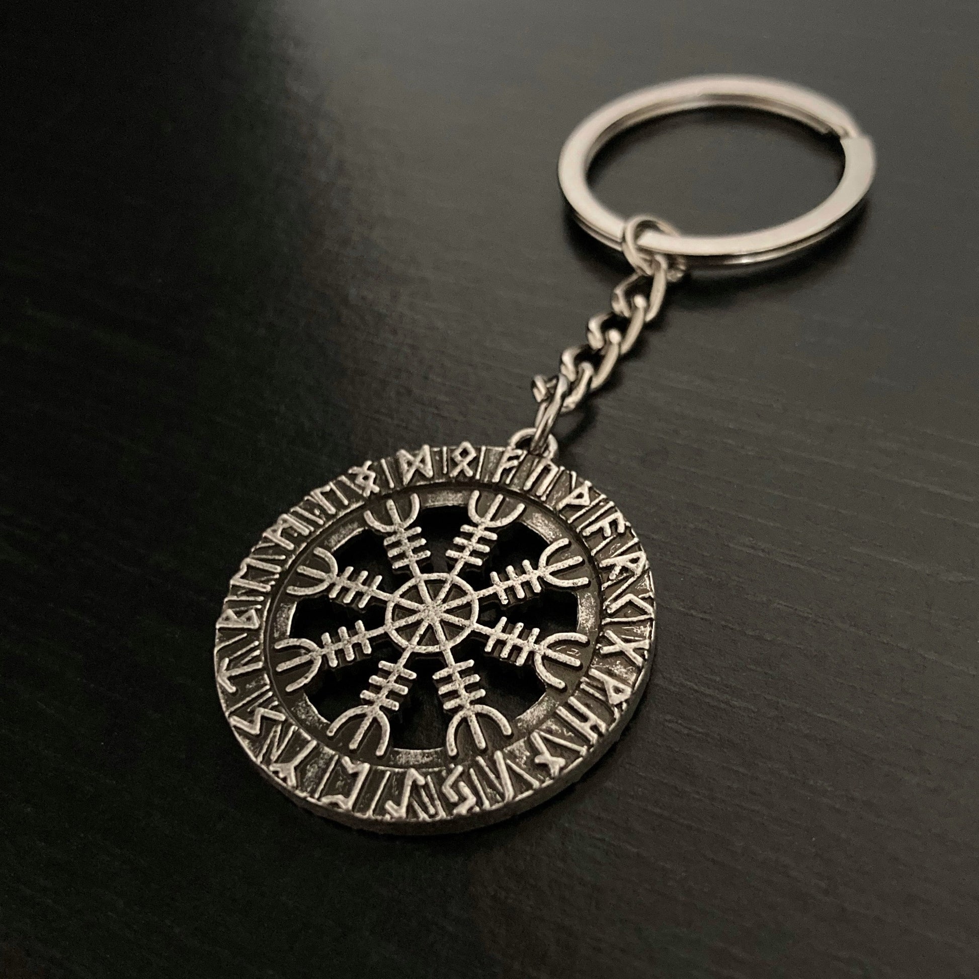 An angled view of a keyring which is round and has a cut out design of the helm of awe symbol in the middle of it. Etched around the outer ring are runes. It is solid to hold but not heavy and the ring part is a shiny silver colour.