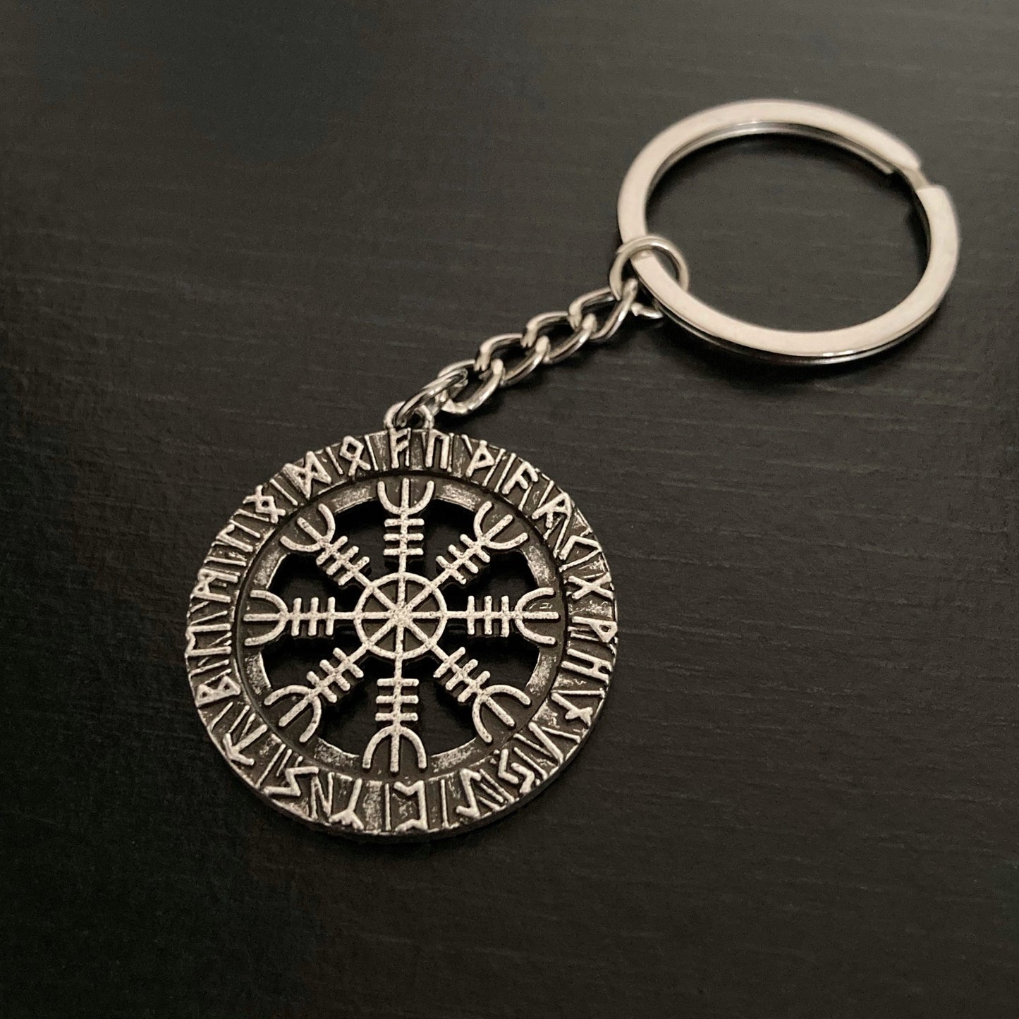 A keyring with intricate detailing on it of both a helm of awe design as well as rune symbols. Between the ring and the main part is a small link chain. All of it is silver in colour.