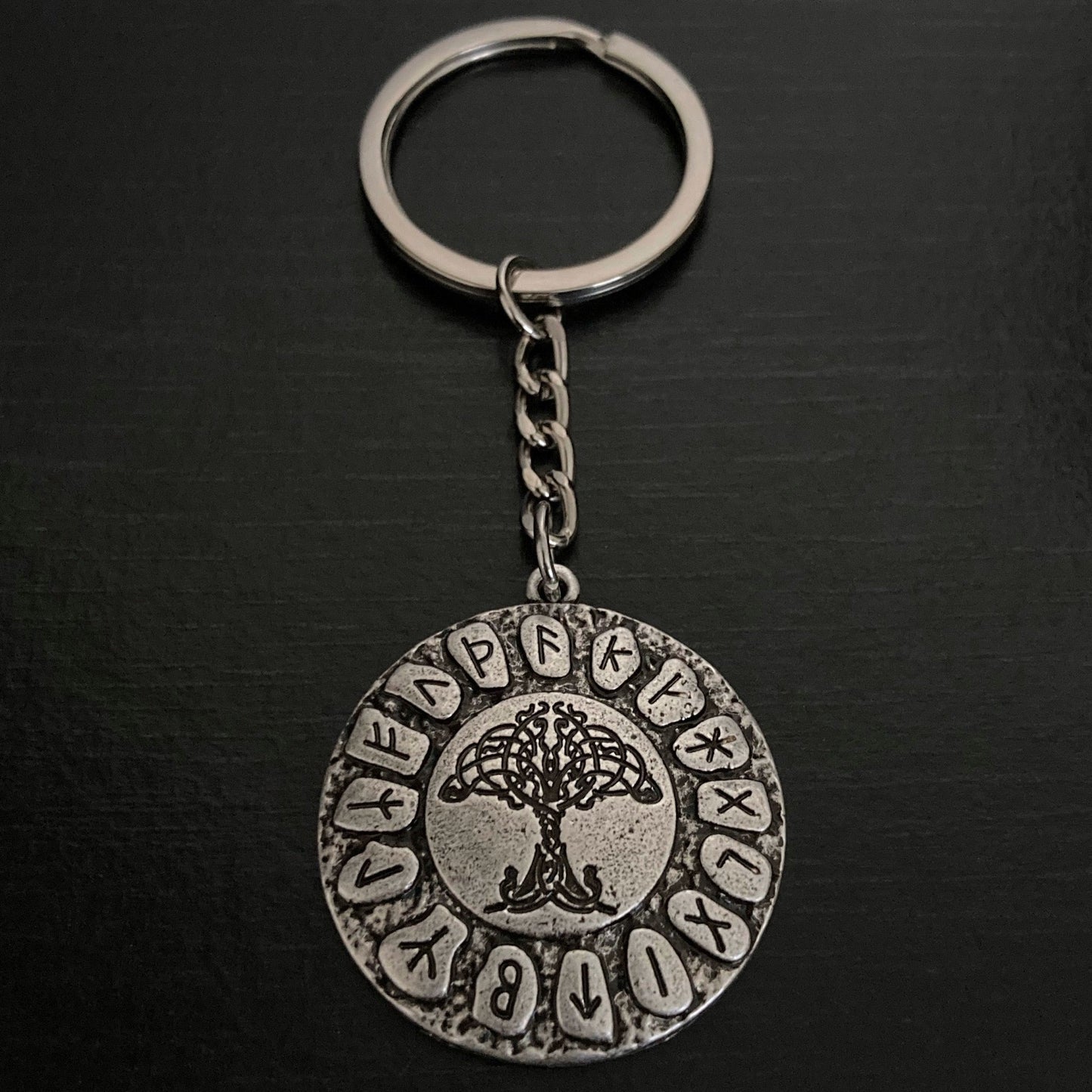 A silver coloured keyring sits on a dark surface. A ring is attached to a small link chain and then there is a circular shape that has runes printed on it around the edge and a tree of life in the centre.