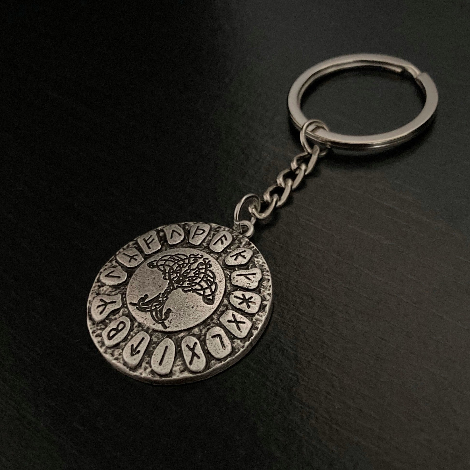 An angled view of a keyring. The main part is a solid circular piece with runes and a tree of life engraved onto it. This is a rustic looking item as opposed to shiny and is light to hold in the hand.