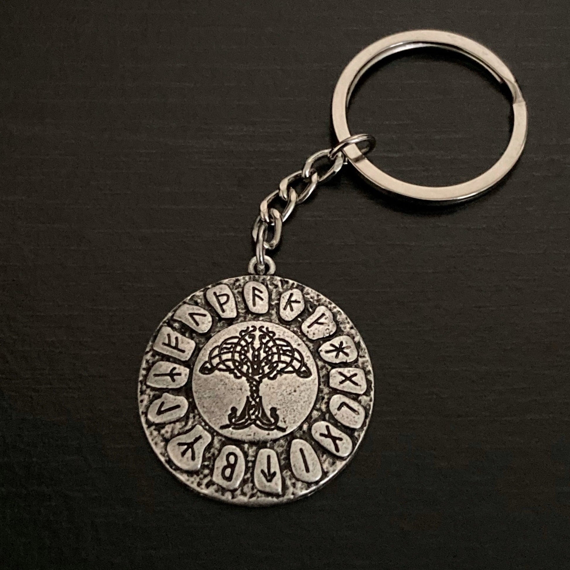 A round coin shaped piece of stainless steel is attached to a chain and ring. In the middle of it a tree of life is seen in black and all around the edge are runic symbols, each in their own oval shaped disc. 