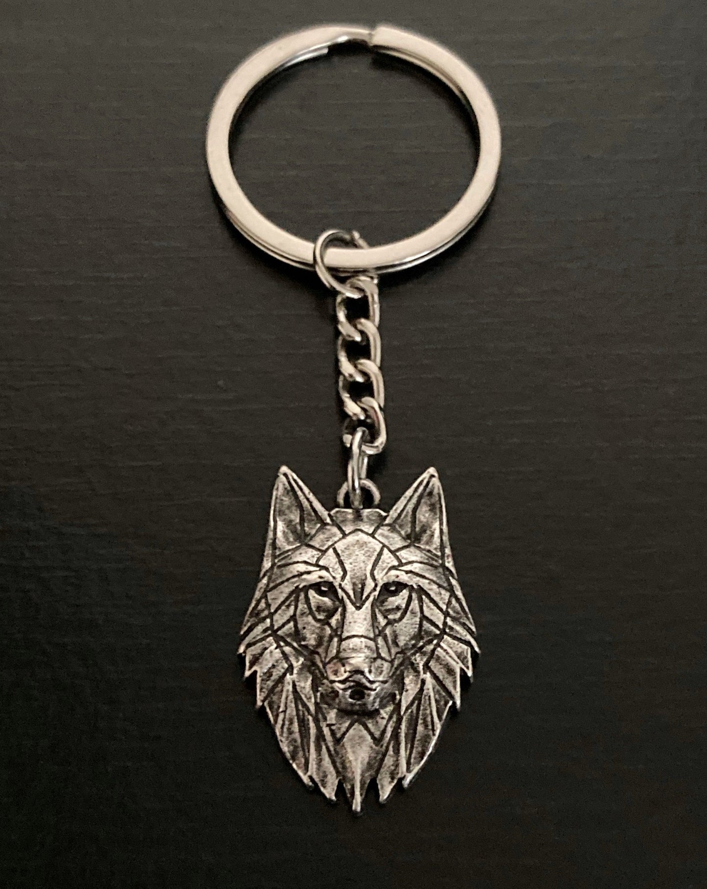 On a black background sits a keyring with a wolfs head pendant hanging from it. It is silver in colour and the wolfs features are visible to see. There is a small link chain seperating the wolf from the ring.