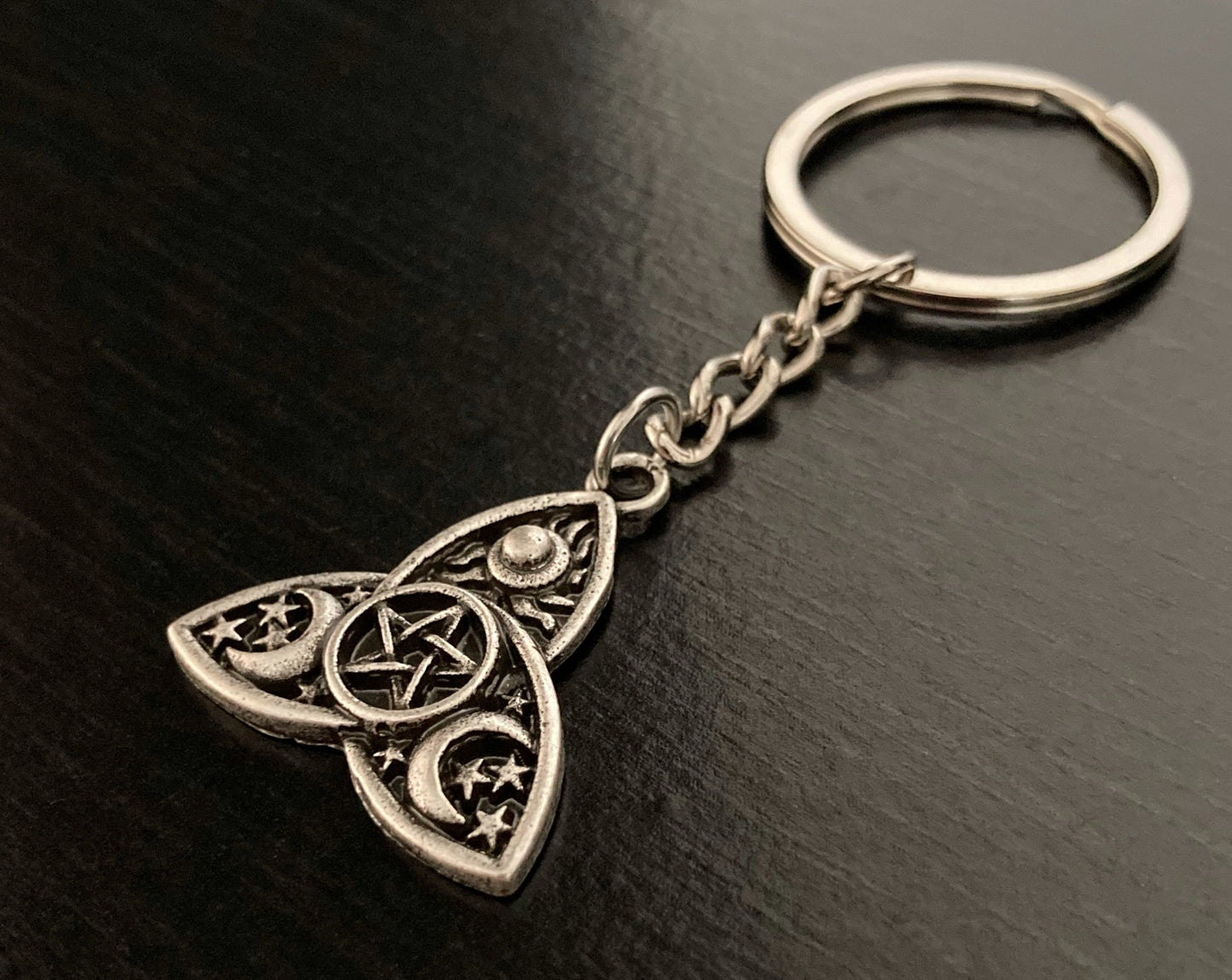 A close up view of a keyring in the shape of triple moons with engravings of moons, stars and a pentagram on it. Silver in colour with a ring for attaching keys to it.