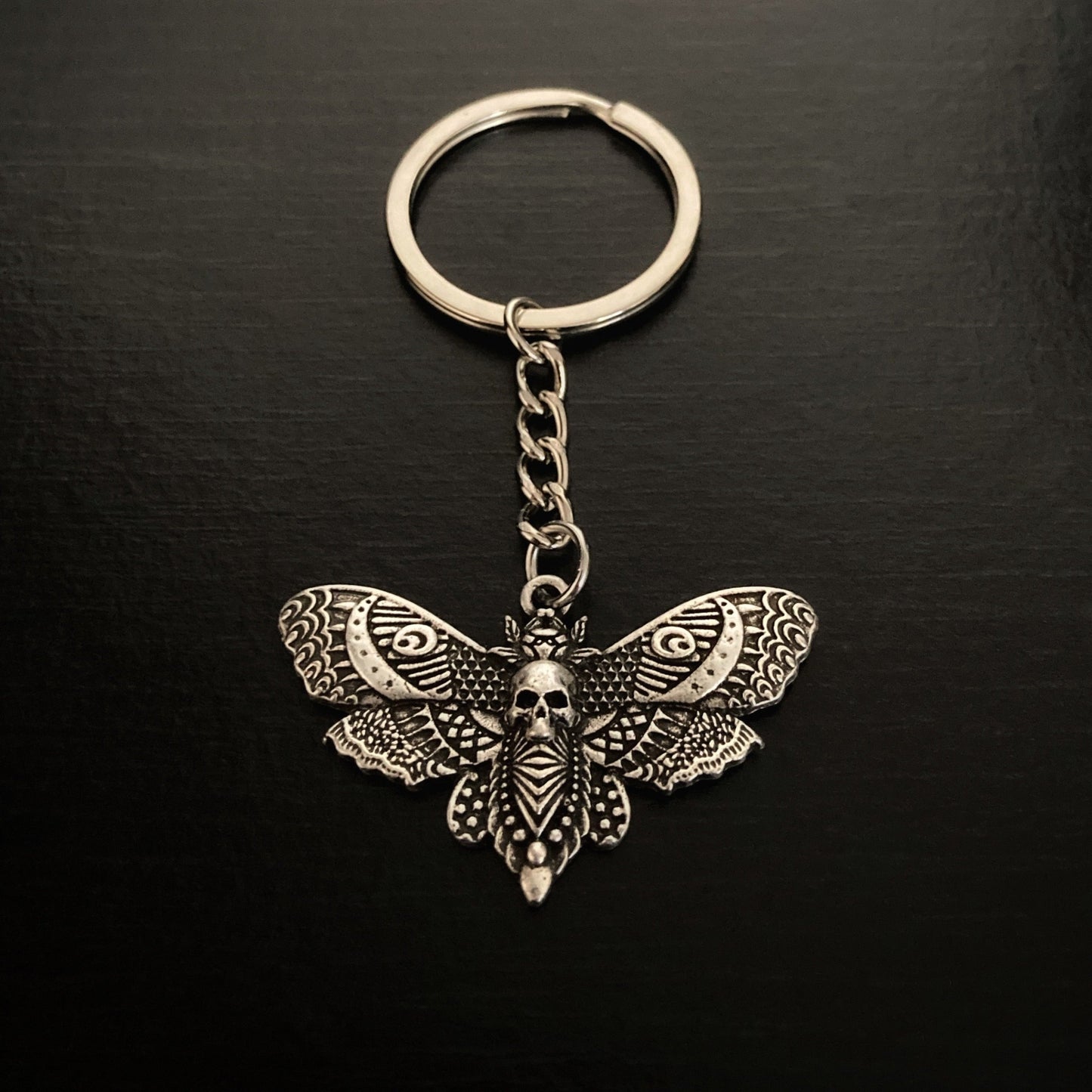 A keyring that has a death moth hanging from it. A small link chain separates the moth from the actual ring. There is very detailed markings on both the moths wings and body with a small skull carving in the middle of it.