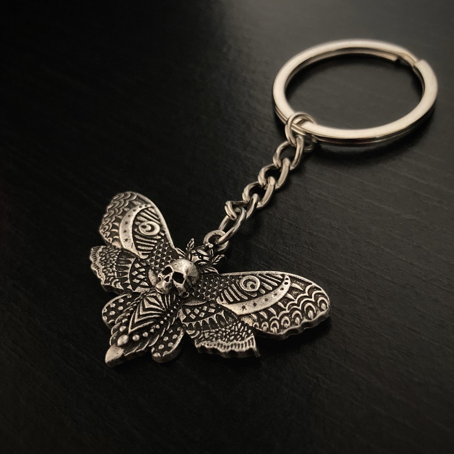 An angled view of a keyring with a death moth pendant attached to it. It is silver in colour and there are beautiful markings all over the moth itself. which you can feel when you touch it.