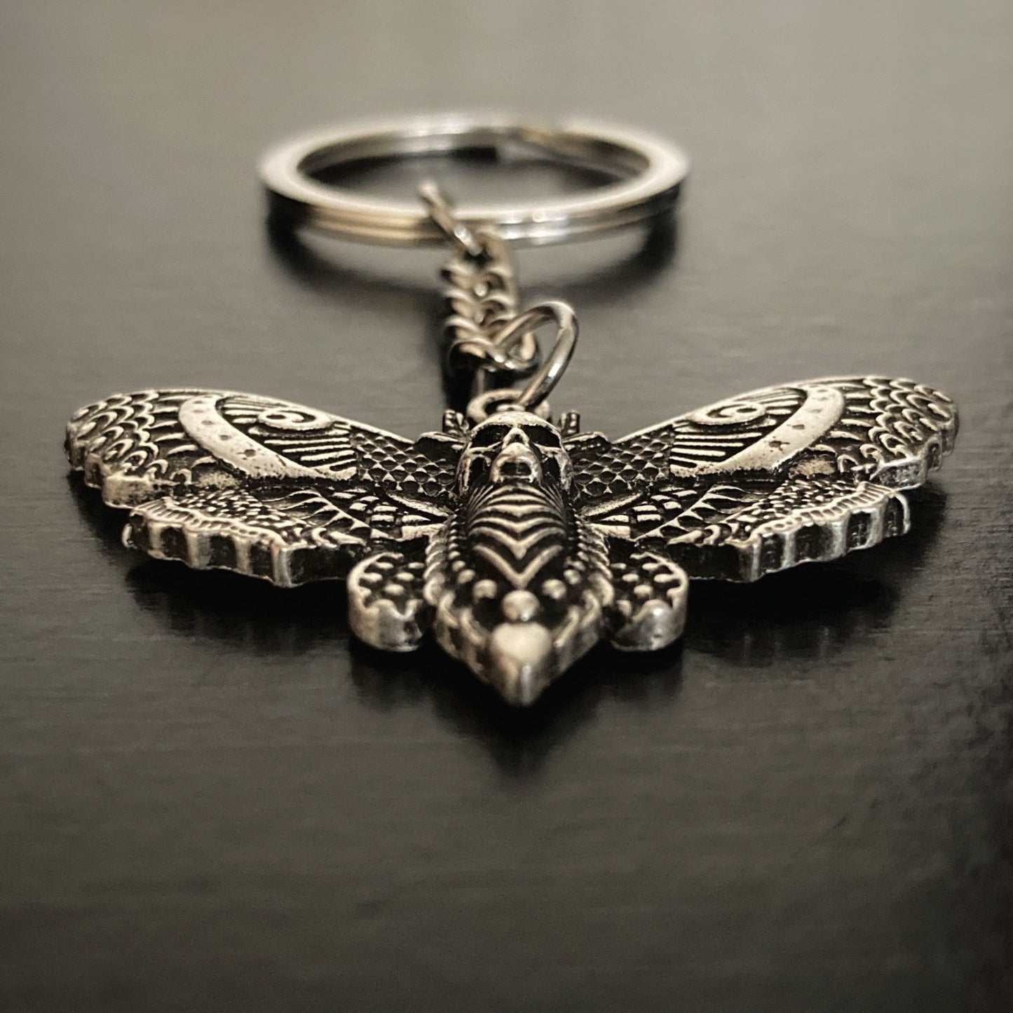 On a black surface lies a stainless steel death moth keyring with a small chain seperating the ring part from the moth itself. The trunk of the moths body has a skull carved into it and the wings have intricate patterning.