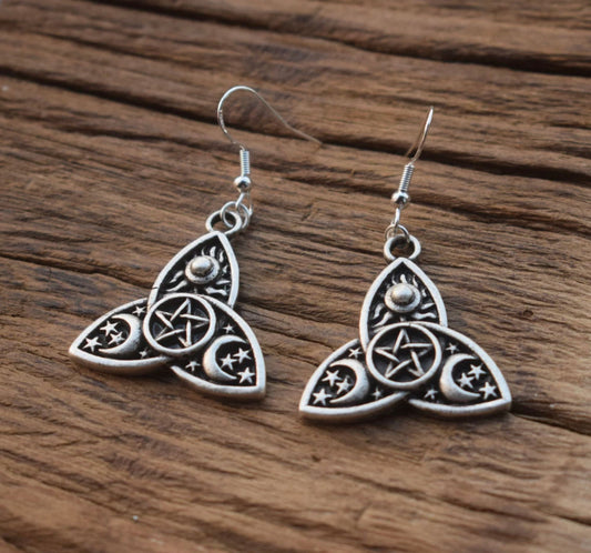 A pair of earrings sit on a dark surface. These are in the shape of a triple moon design with a pentagram etched into a small circle in the middle of this. There are also moons and stars etched onto the outer part of the design.