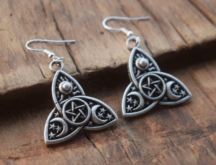 Two silver coloured earrings lean against some wood. There is a hook for putting in your ear and the earring design is intricate in its patterning.