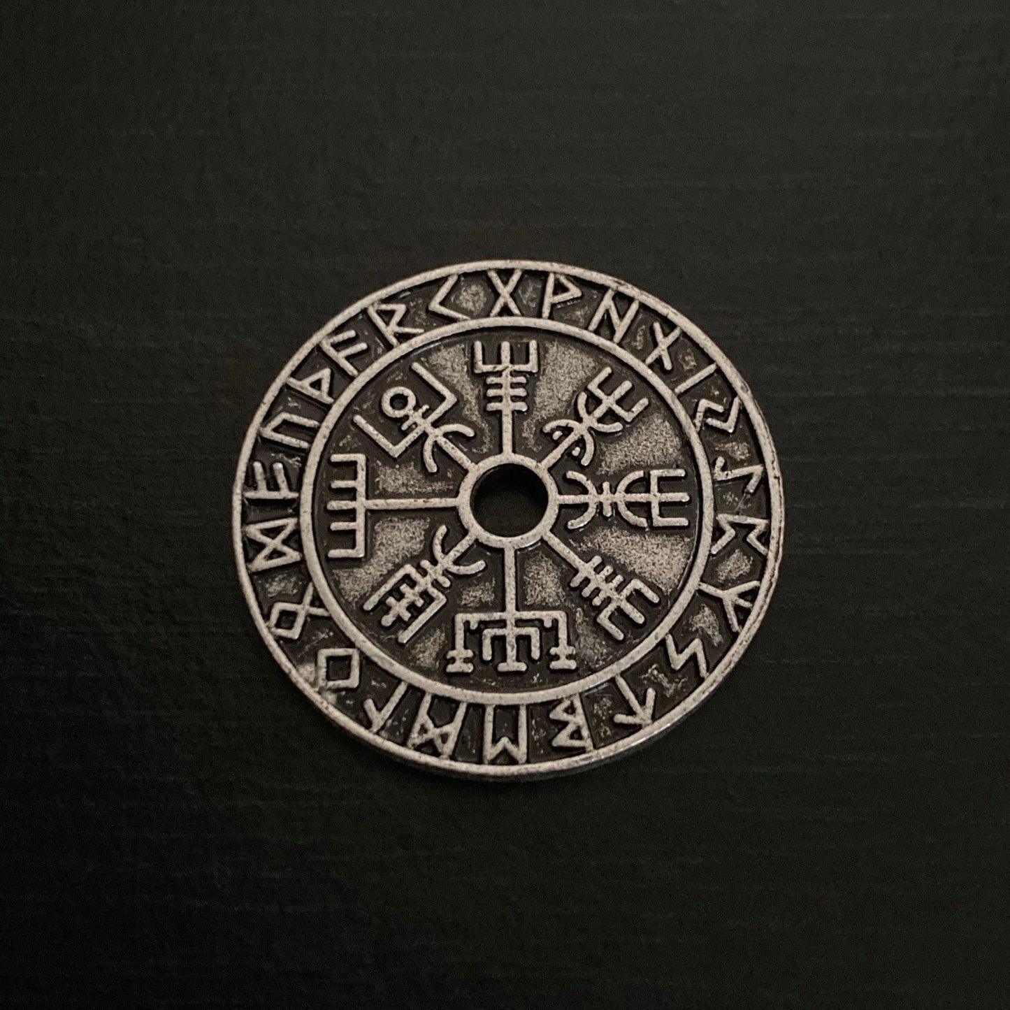 A round coin is on a black background. It is a rustic looking and features a hole in the centre of it with a vegvisir design coming away from that an outer circle of rune symbols all around the edge.