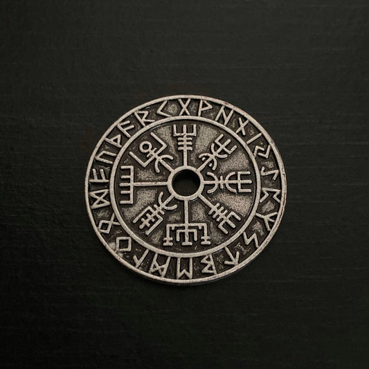 A round coin is on a black background. It is a rustic looking and features a hole in the centre of it with a vegvisir design coming away from that an outer circle of rune symbols all around the edge.