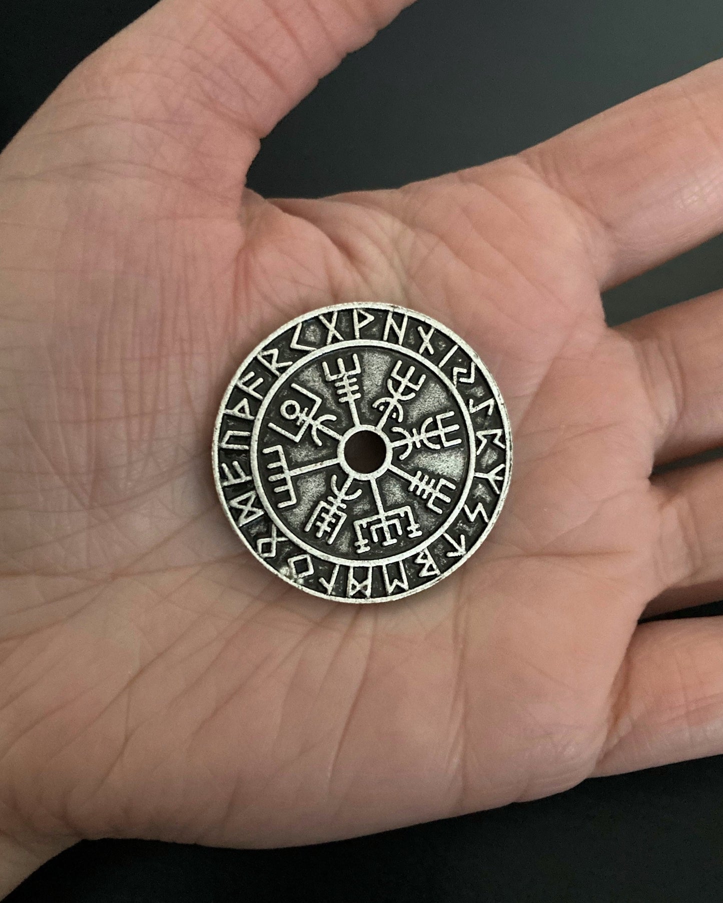 A round coin sits in the open palm of a hand. It features a hole in the middle of it with a vegvisir design coming off of that. There is then an outer ring that has runes engraved all around it.