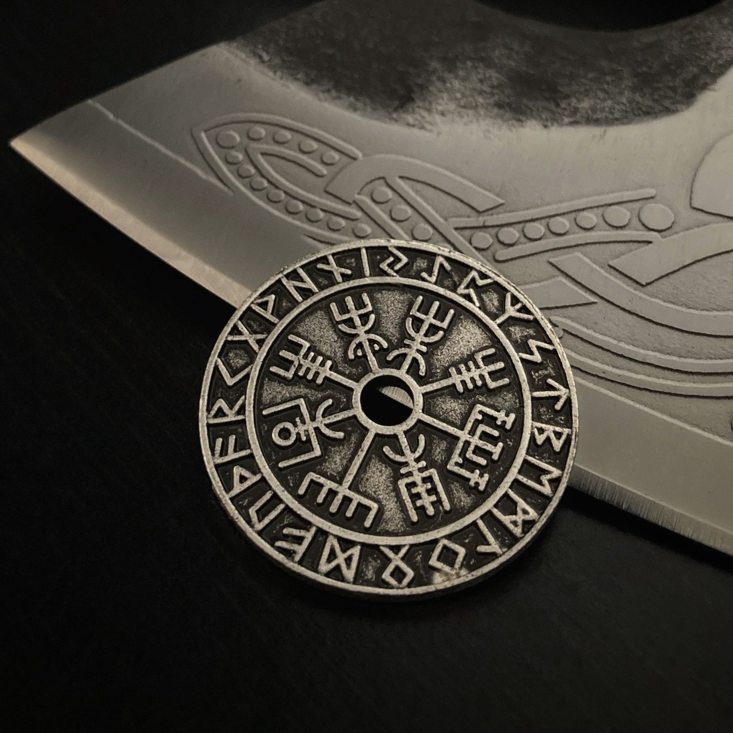 Leaning against a axe blade sits a viking style coin. Circular in shape with a hole in the middle. A vegvisir is etched around the hole and an outer ring of runic symbols goes around the outside