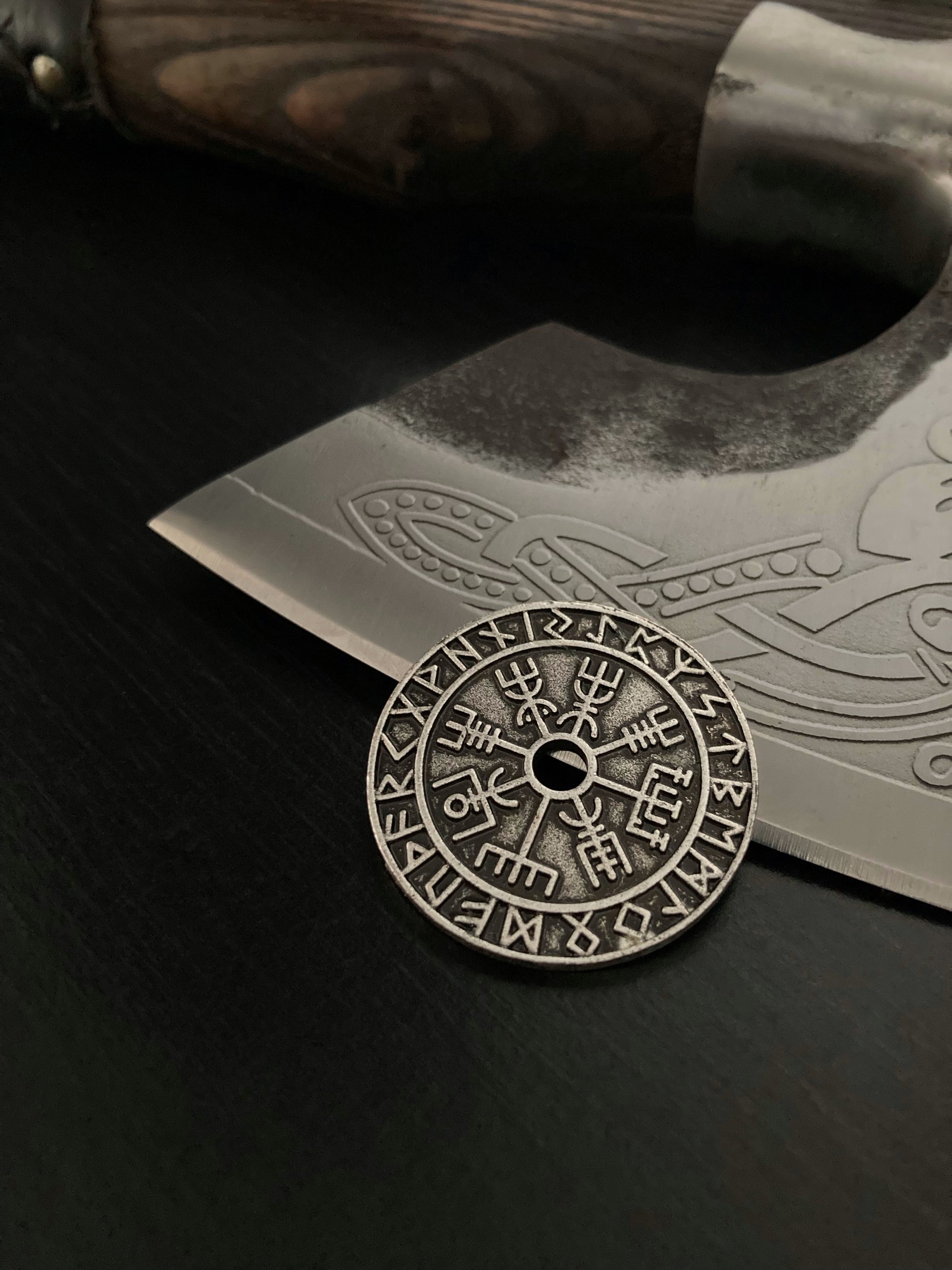 Leaning against a axe blade sits a viking style coin. Circular in shape with a hole in the middle. A vegvisir is etched around the hole and an outer ring of runic symbols goes around the outside.