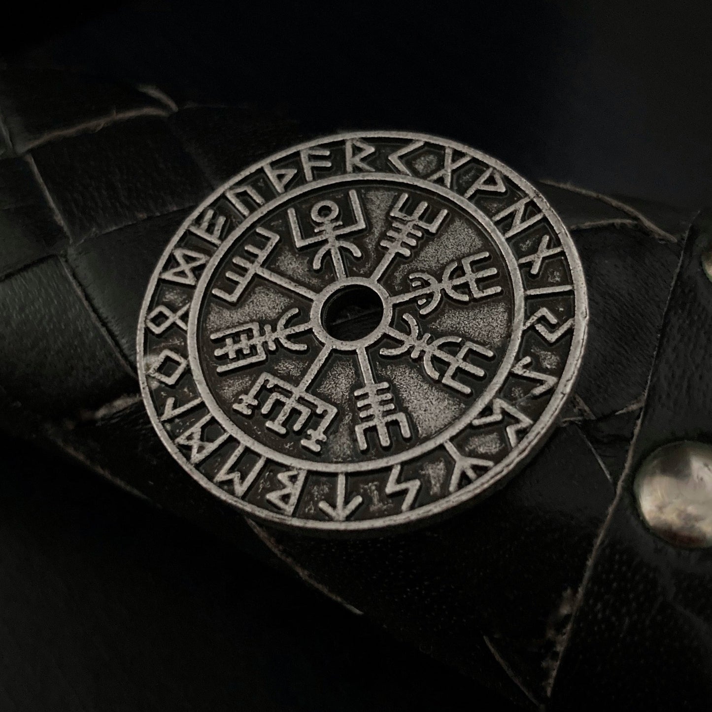 On a patterned black surface sits a round coin that is both solid and weighty. With a vegvisir and rune design this coin has a rustic look to it rather than a shiny one.