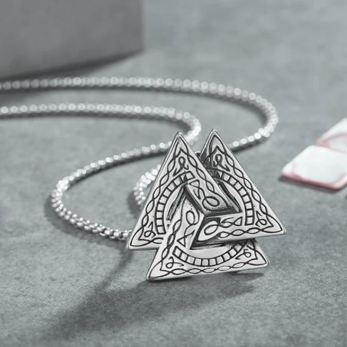 On a grey surface is a silver coloured chain attached to a pendant the same colour which is 3 interlocking tiangles. A patterned marking covers the pendant.