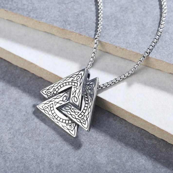 On a grey background lies a triangular shaped pendant with black etched markings on it. This item is in the style of a viking valknut and is attached to a sturdy looking chain, both are silver in colour.