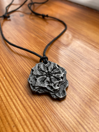 Lying on a wooden surface is a black corded necklace with a pendant in the shape of a rock attached to it. The item has a helm of awe engraved onto the top of it in black and is weighty. It has several jagged but not sharp edges to it.